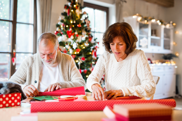 Beautiful senior couple in white woolen sweaters sitting at the table wrapping Christmas gifts together.