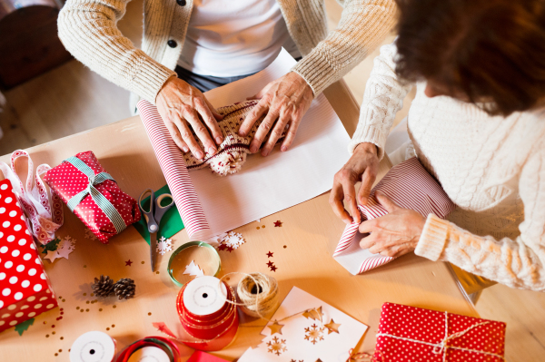 Unrecognizable senior couple in white woolen sweaters sitting at the table wrapping Christmas gifts together. Aerial view.