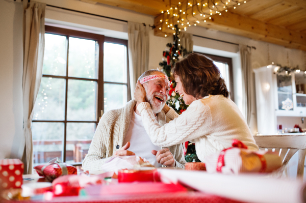 Beautiful senior couple in white woolen sweaters sitting at the table wrapping Christmas gifts together, having fun.