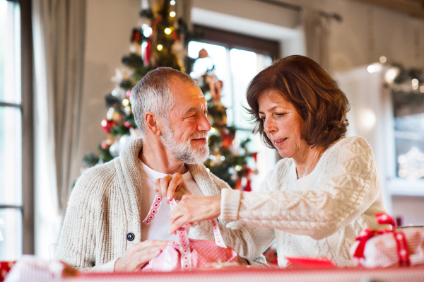 Beautiful senior couple in white woolen sweaters sitting at the table wrapping Christmas gifts together.