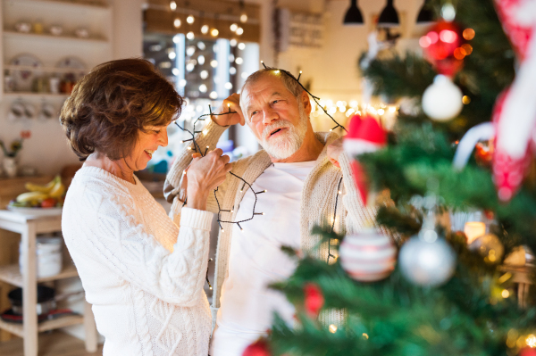 Beautiful senior couple in white woolen sweaters decorating Christmas tree inside their house. Man and woman having fun.