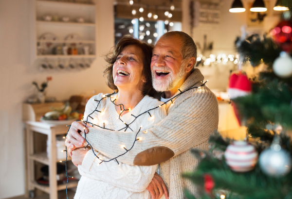 Beautiful senior couple in white woolen sweaters hugging at home. Man and woman having fun at Christmas time.