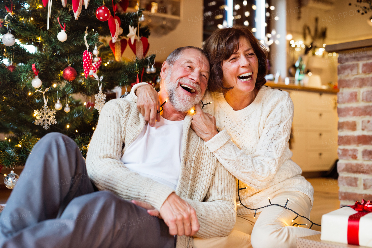 Senior couple sitting on the floor in front of illuminated Christmas tree inside their house, talking and laughing. Man entangled in chain of lights.