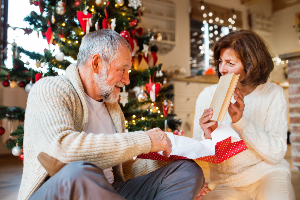 Senior couple sitting on the floor in front of illuminated Christmas tree inside their house giving presents to each other, woman unwrapping her gift.