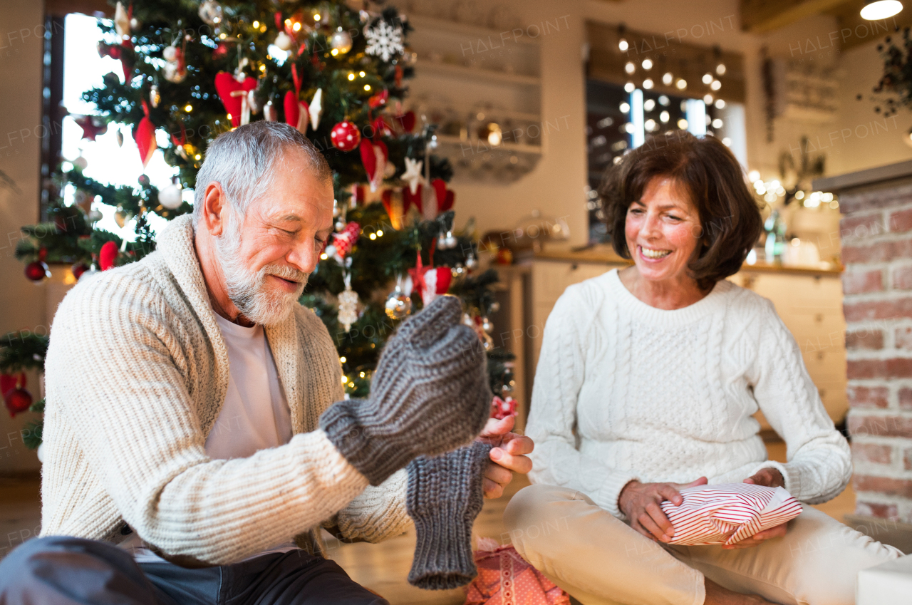 Senior man sitting on the floor in front of illuminated Christmas tree with his wife. The man is trying on knitted mittens.