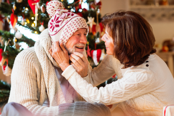 Senior man sitting on the floor in front of illuminated Christmas tree with his wife. The man is trying on knitted scarf, hat and mittens.