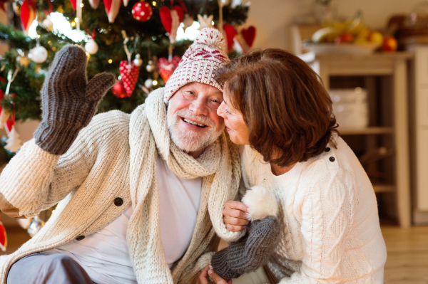 Senior man sitting on the floor in front of illuminated Christmas tree with his wife. The man is trying on knitted scarf, hat and mittens.