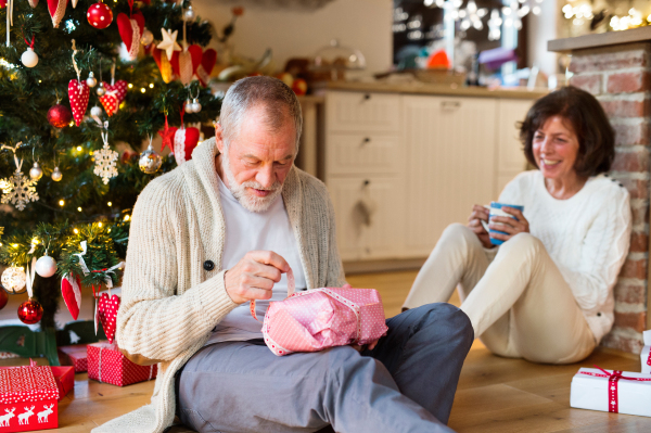 Senior couple sitting on the floor in front of illuminated Christmas tree inside their house. Man unpacking a present.