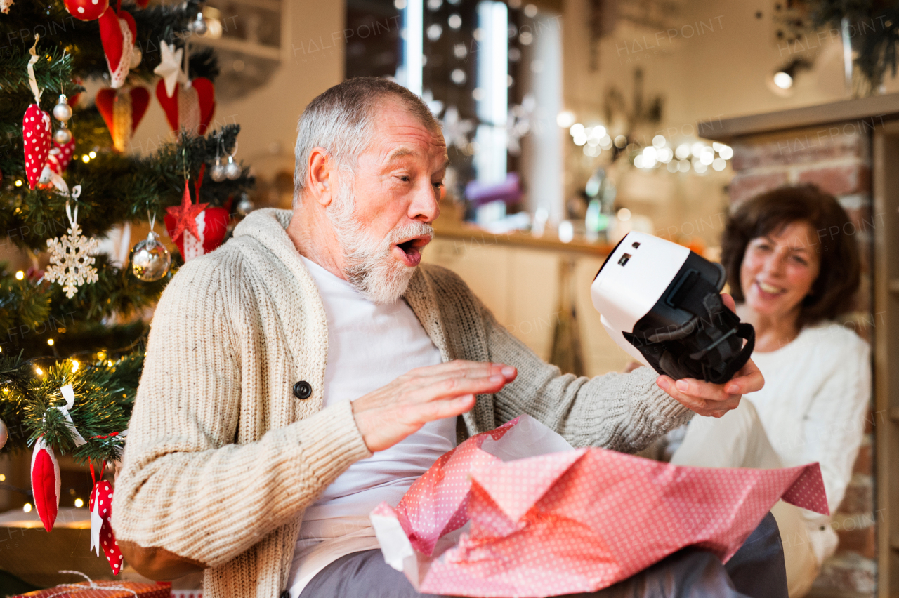 Senior couple sitting on the floor in front of illuminated Christmas tree inside their house unpacking VR glasses.