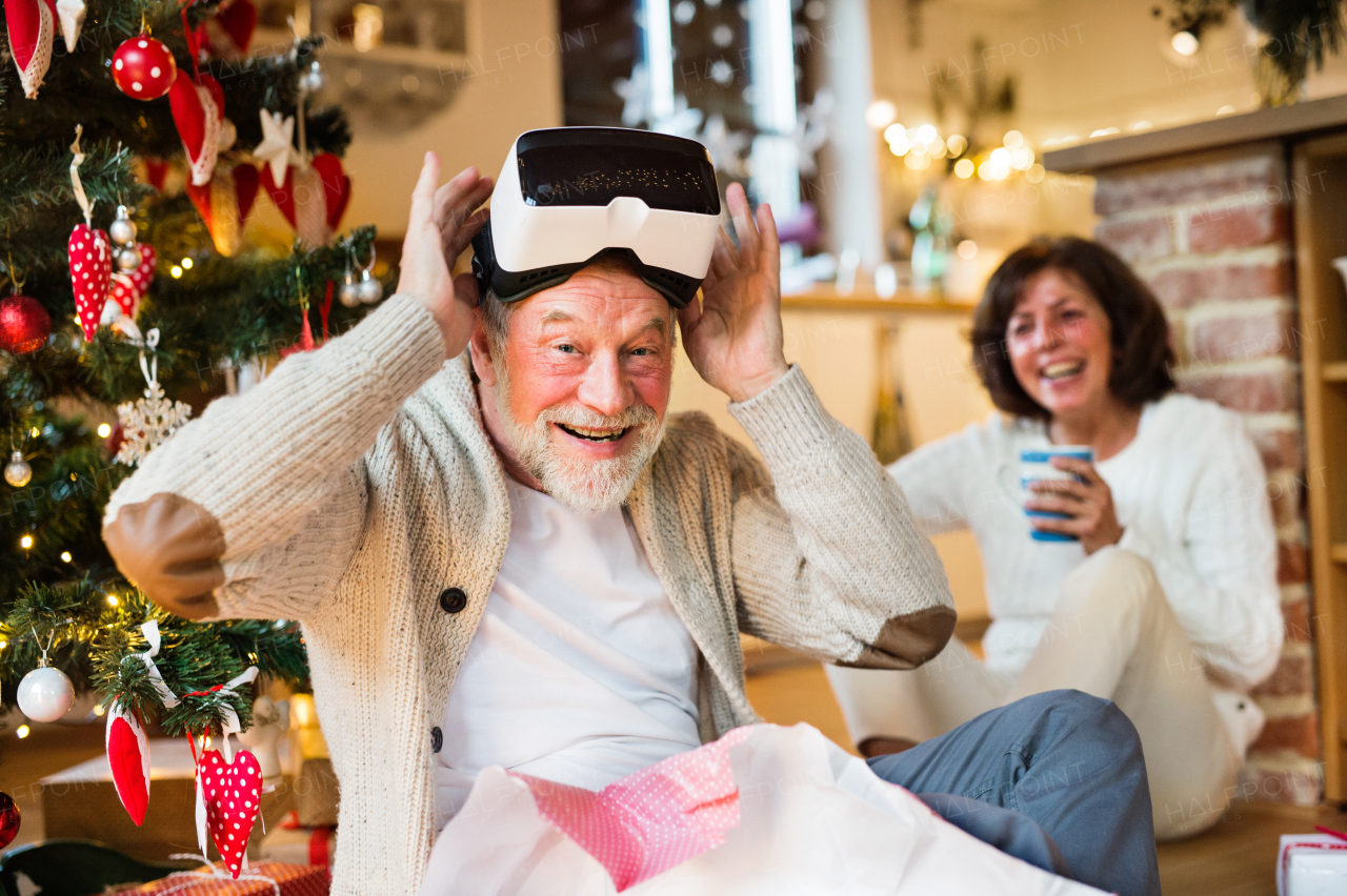 Senior couple sitting on the floor in front of illuminated Christmas tree inside their house unpacking and trying on VR glasses.