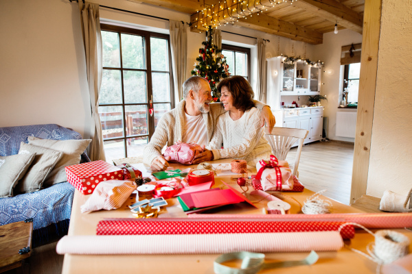 Beautiful senior couple in white woolen sweaters sitting at the table wrapping Christmas gifts together.