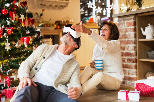 Senior couple sitting on the floor in front of illuminated Christmas tree inside their house unpacking and trying on VR glasses.