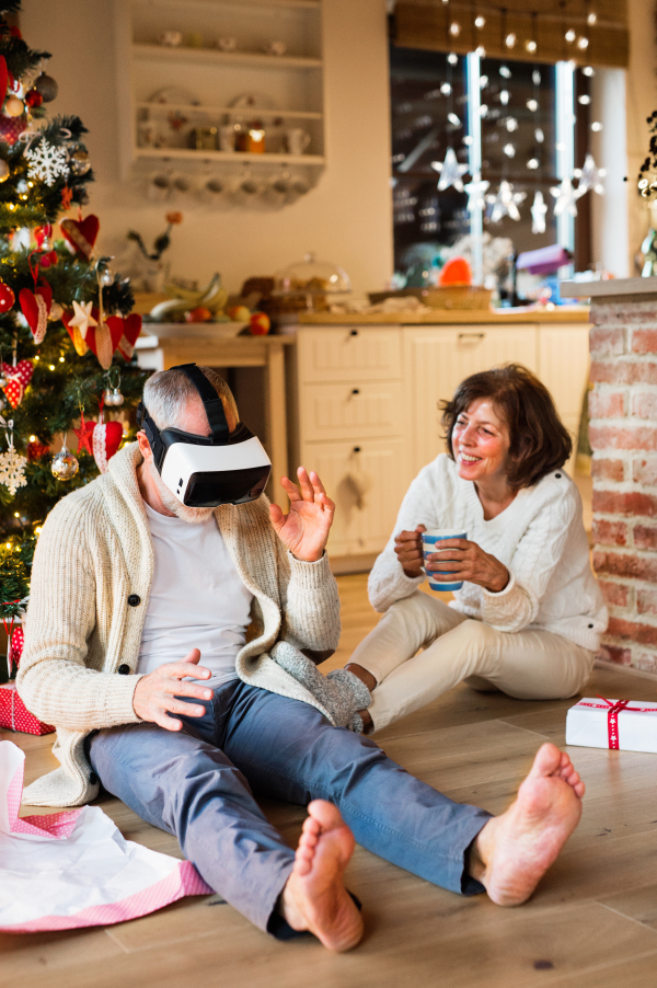 Senior couple sitting on the floor in front of illuminated Christmas tree inside their house unpacking and trying on VR glasses.