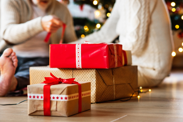 Unrecognizable senior couple sitting on the floor in front of illuminated Christmas tree inside their house giving presents to each other.