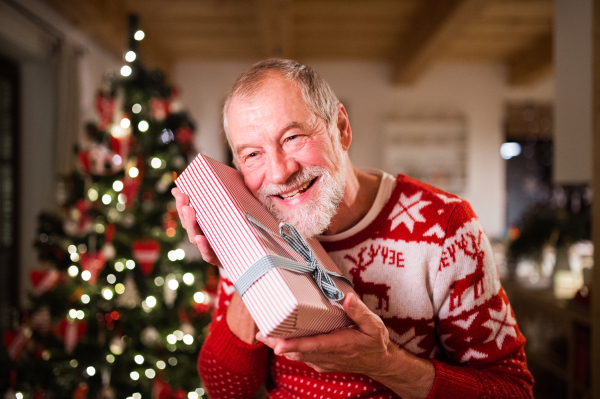 Senior man standing in front of illuminated Christmas tree inside his house holding a present, feeling pleased.