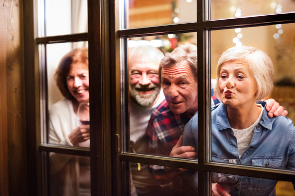 Senior friends with wine glasses at Christmas time. Men and women looking out of the window, having fun. Shot through glass.