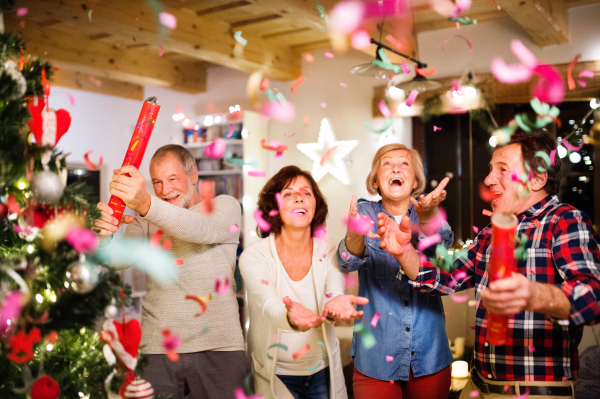 Senior friends with confetti poppers next to Christmas tree, having fun.