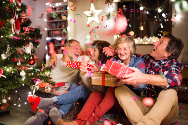 Senior friends sitting on the wooden floor next to the illuminated Christmas tree with their Christmas presents, talking.