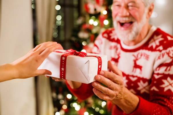 Senior man standing in front of illuminated Christmas tree inside his house holding a present, laughing.