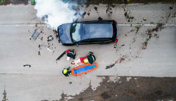 Three unrecognizable firefighters helping a young injured woman lying on the road after an accident. Top view.