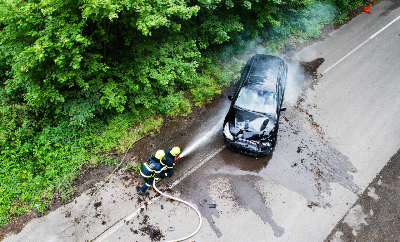 Aerial view of two firefighters extinguishing a burning car after an accident on the road in the countryside.