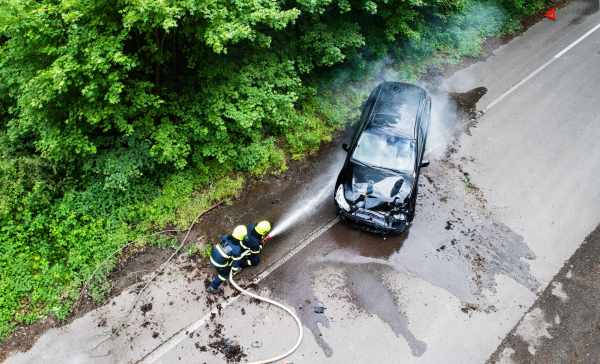 Aerial view of two firefighters extinguishing a burning car after an accident on the road in the countryside.