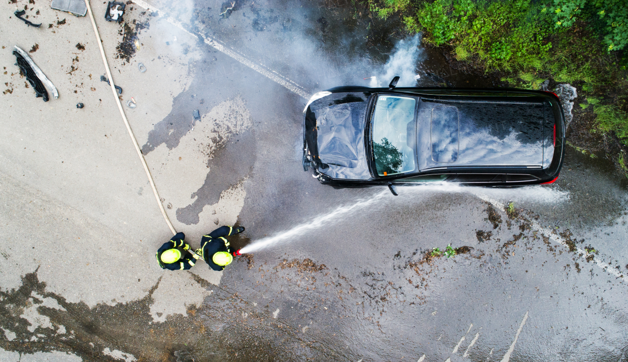 Two firefighters extinguishing a burning car after an accident on the road in the countryside. Top view.