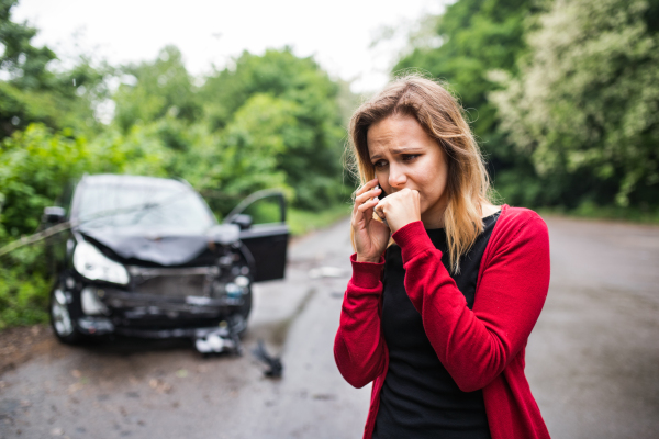 A frustrated young woman with smartphone by the damaged car after a car accident, making a phone call.