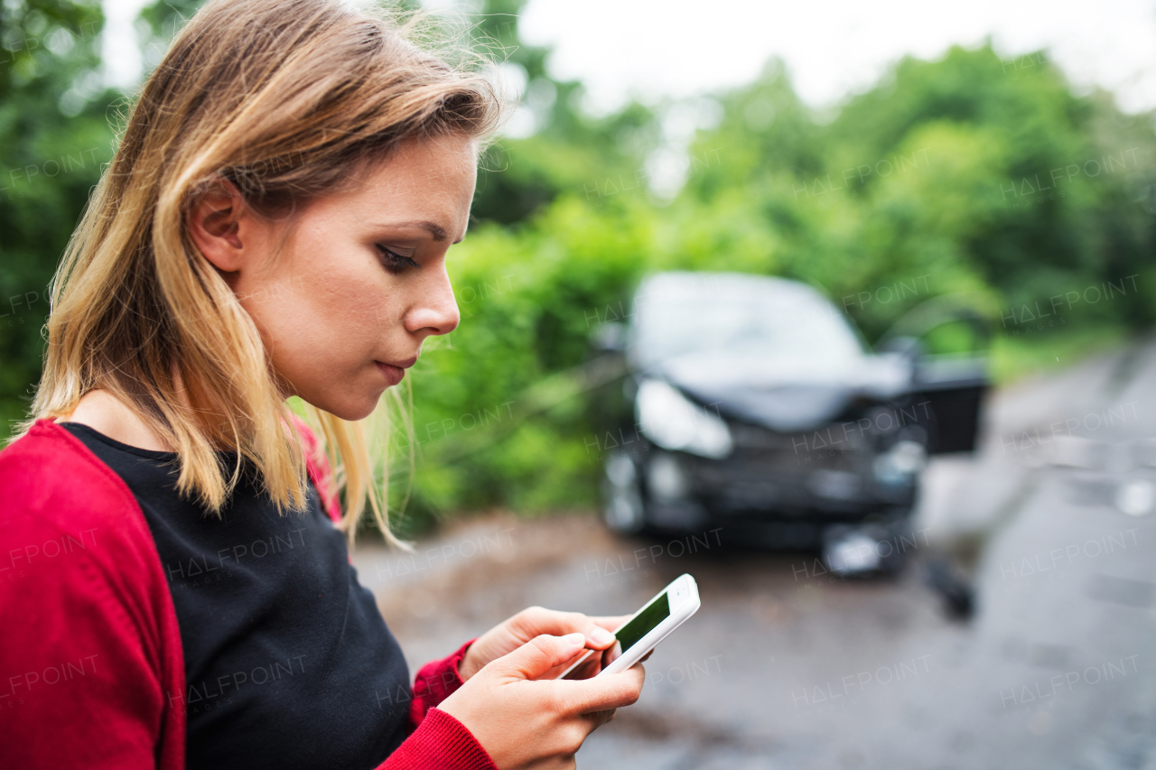 A close-up of a young woman with smartphone by the damaged car after a car accident, text messaging. Copy space.