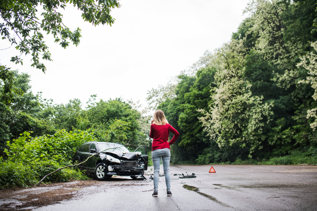 A young woman with smartphone by the damaged car after a car accident, making a phone call. Rear view.