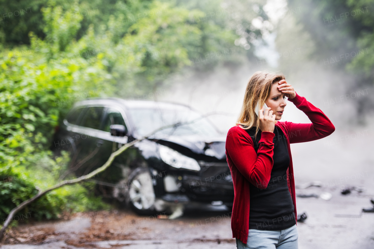 A frustrated young woman with smartphone by the damaged car after a car accident, making a phone call. Copy space.