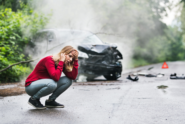 A frustrated young woman with smartphone by the damaged car after a car accident, making a phone call. Copy space.