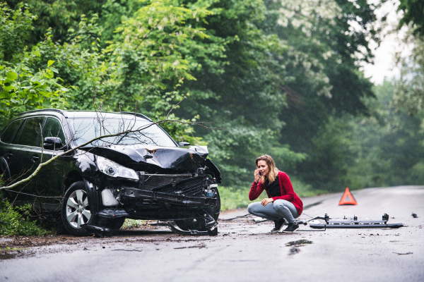 A frustrated young woman with smartphone by the damaged car after a car accident, making a phone call. Copy space.