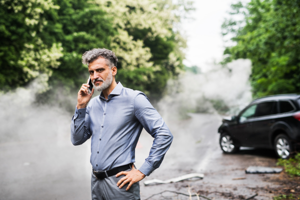 Handsome mature man making a phone call after a car accident, smoke in the background. Copy space.