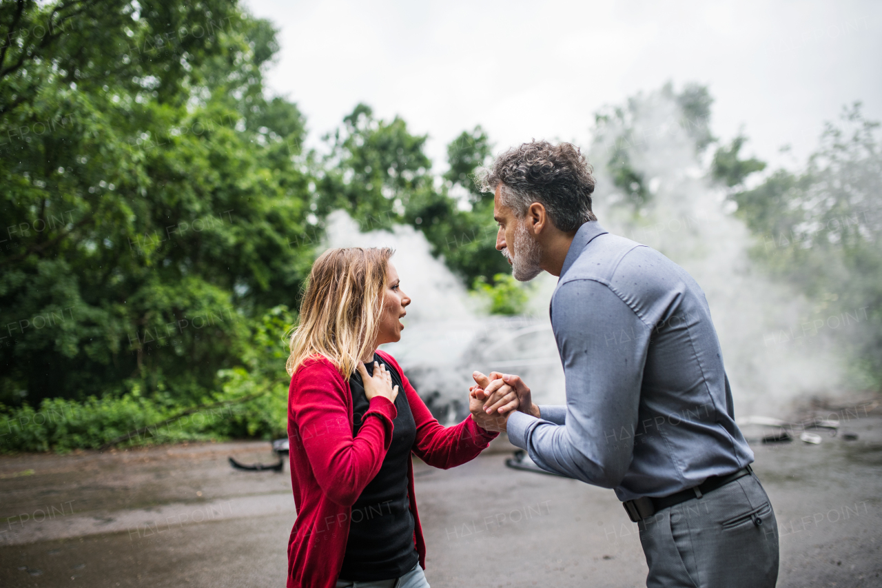 A mature man talking to a stressed young woman after a car accident, helping her. A smoke coming from a broken car in the background.