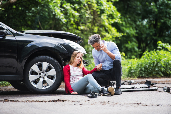 Young woman by the car after an accident and a man with smartphone, making a phone call.