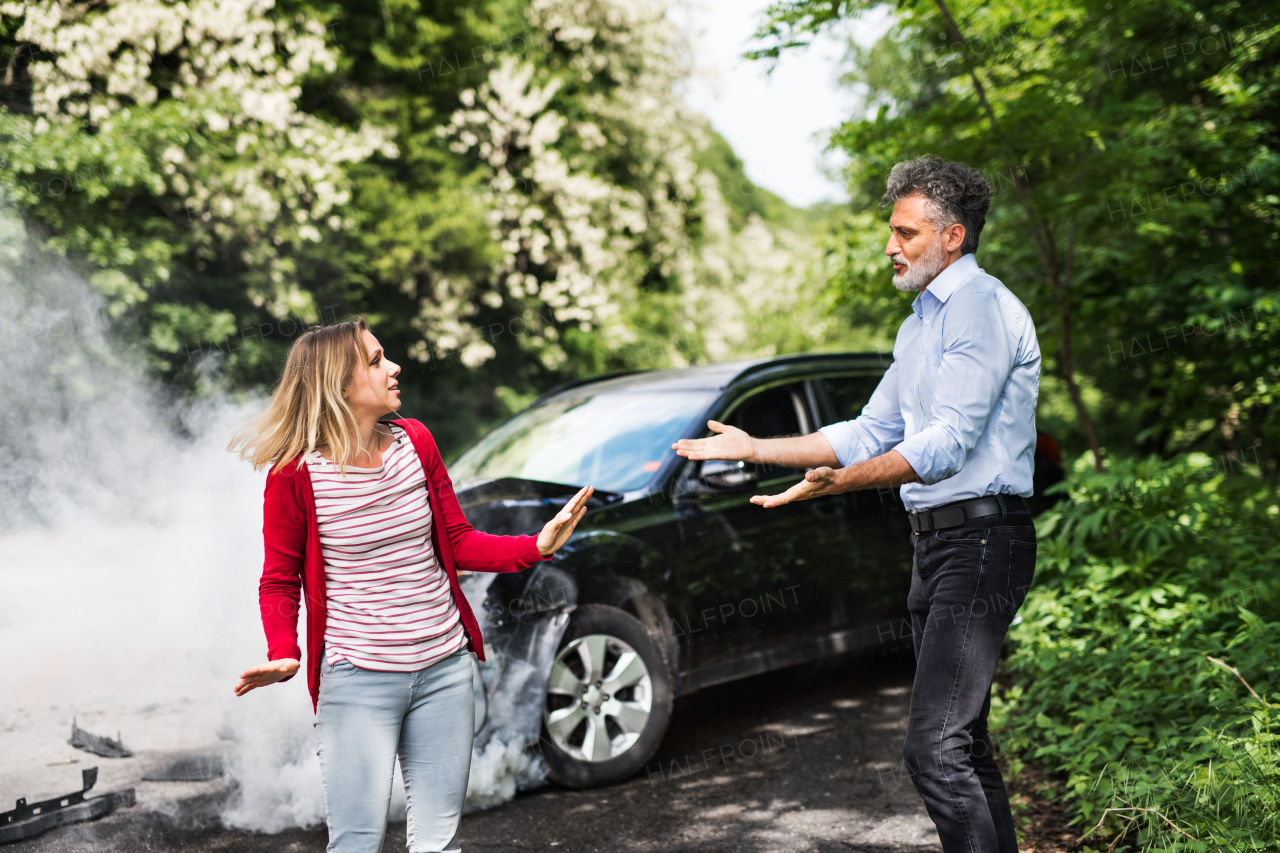 An angry mature man and young woman arguing after a car crash. A smoke coming from a broken car in the background.