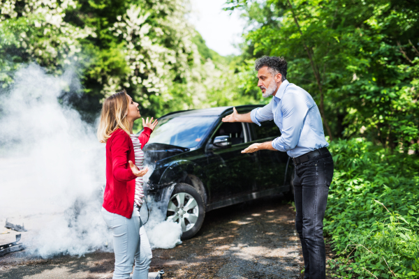 An angry mature man and young woman arguing after a car crash. A smoke coming from a broken car in the background.