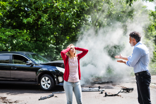 An angry mature man and young woman arguing after a car crash. A smoke coming from a broken car in the background.