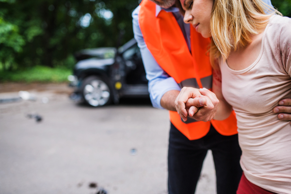 An unrecognizable man helping a young injured woman to walk away from the car after an accident.