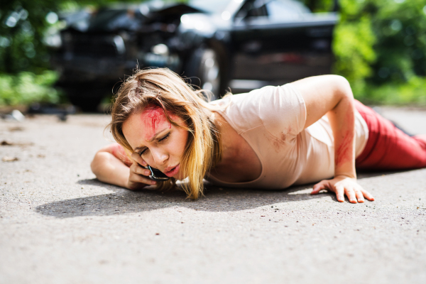 Young injured woman lying on the road in front of a damaged car after a car accident, making a phone call.