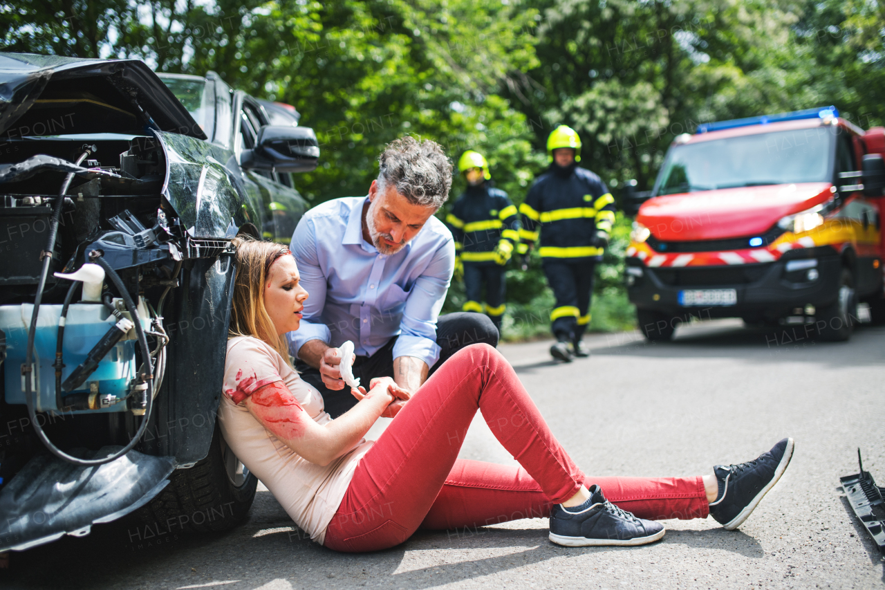 A man helping a young woman on the road after a car accident, firefighters in the background.