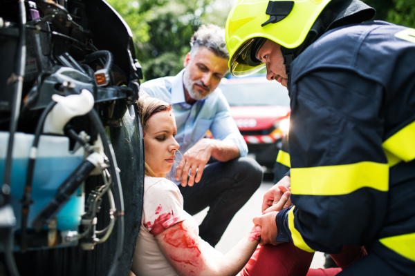 A firefighter helping a young injured woman sitting by the car on a road after an accident, unconscious.