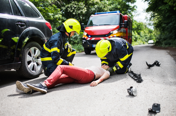 Firefighters helping a young woman after a car accident. A female driver lying down on the countryside road.