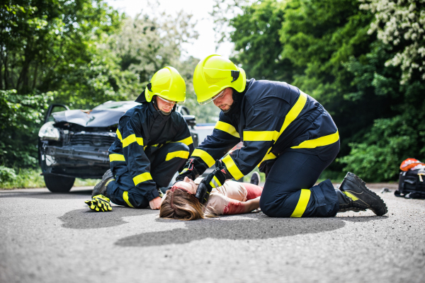 Firefighters helping a young woman after a car accident. A female driver lying down on the countryside road.