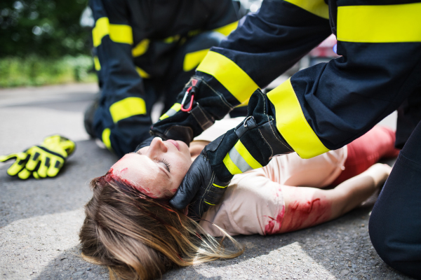 Unrecognizable firefighters helping a young injured woman lying on the road after an accident, unconscious.