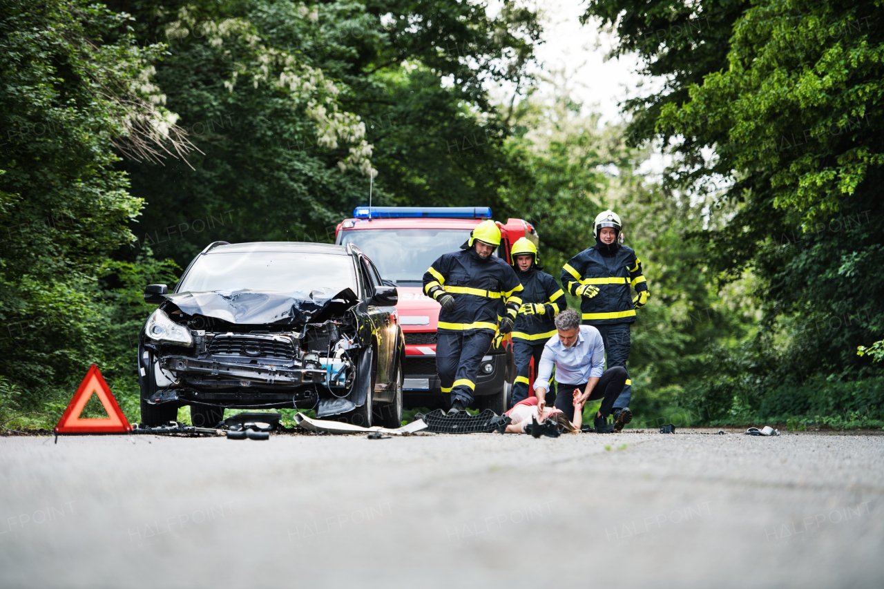 Firefighters running to rescue a woman lying unconscious on the road after a car accident. A mature man holding her hand, checking her pulse. Copy space.