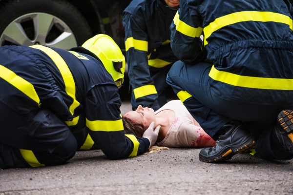 Three unrecognizable firefighters helping a young injured woman lying on the road after an accident.