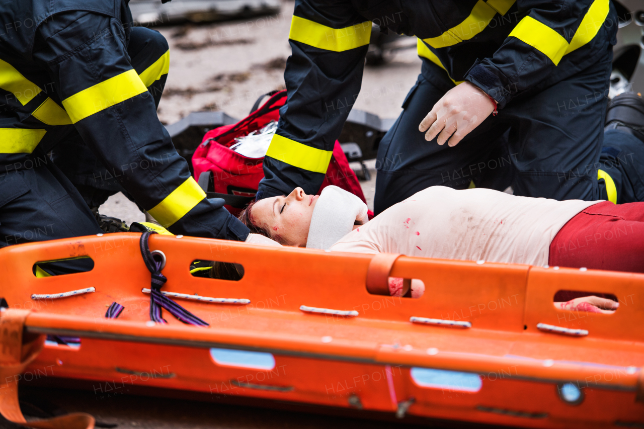 Unrecognizable firefighters helping a young injured woman lying on the road after an accident. A plastic stretcher in the foreground.