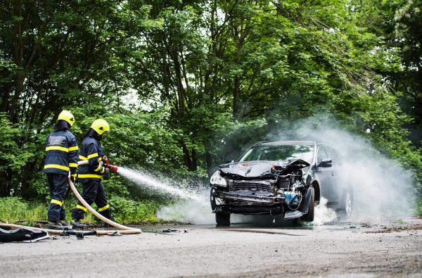 Two firefighters extinguishing a burning car after an accident on the road in the countryside.